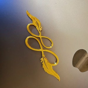 Könnte beinhalten: Goldfarbenes Metallsymbol für Unendlichkeit mit zwei Federdesigns an jedem Ende.