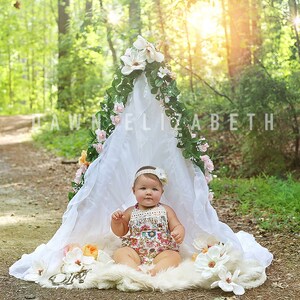 Könnte beinhalten: Ein kleines Mädchen in einem Blumenkleid sitzt in einem weißen Stoff-Tipi, das mit Grünpflanzen und Blumen dekoriert ist. Das Tipi steht in einem Waldgebiet mit einem unbefestigten Weg, der vom Tipi wegführt.