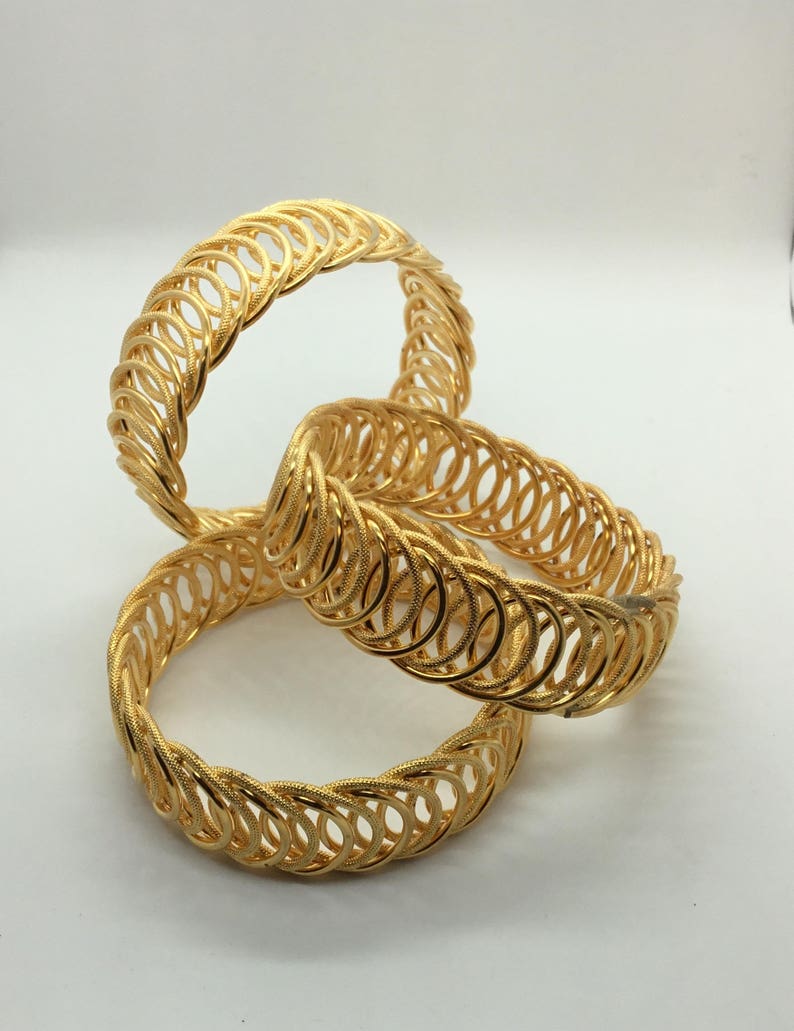 May include: Three gold bangles with a woven design. The bangles are stacked on top of each other.