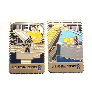 May include: Two rectangular metal enamel pins designed to resemble postage stamps. Each pin features a U.S. Postal Service worker in a sorting facility, with the text U.S. POSTAL SERVICE 8c printed at the bottom in blue and black lettering.