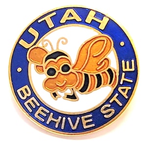 May include: A round pin with a blue border and gold lettering that reads "UTAH BEEHIVE STATE." The center features a cartoon bee in orange, black, and gold. The pin is likely a souvenir or emblem.