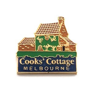 May include: A gold-toned metal enamel pin featuring a depiction of a cottage with brown brick walls, a brown roof, and green ivy. Below the cottage, a dark blue rectangular banner displays the gold text Cooks' Cottage MELBOURNE in capital letters.