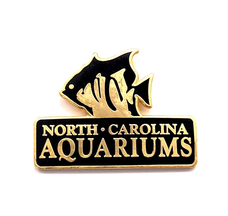 May include: A gold-coloured pin featuring a black fish design atop a black rectangular base. The base reads "NORTH CAROLINA AQUARIUMS" in gold lettering. The pin is likely a souvenir or collectible item.