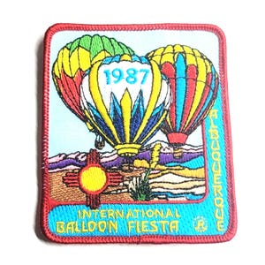 May include: A rectangular embroidered patch featuring three colorful hot air balloons against a light blue sky. The center balloon displays the year 1987. Below, a Zia symbol and desert landscape are depicted with the text Albuquerque International Balloon Fiesta.