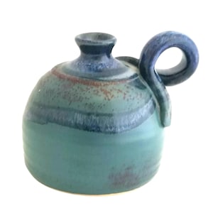 May include: A small, handcrafted ceramic jug with a loop handle. The jug features a rounded body and a dark blue rim. The body is primarily teal with bands of darker blue and brown speckles. The handle is a matching dark blue.
