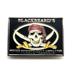 May include: A rectangular metal pin featuring a black background with a central oval frame. Inside the oval is a white skull wearing a red striped bandana, positioned over two crossed swords with yellow hilts. Text reads BLACKBEARD'S and SINCE PUERTO VALLARTA 1768.