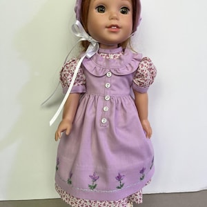 May include: A doll wearing a lavender dress with embroidered flowers, a matching bonnet, and white shoes. The dress has a floral print on the sleeves and hem. A white ribbon is tied around the doll's neck.