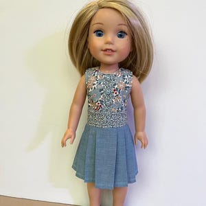 May include: A doll with blonde hair and blue eyes wearing a sleeveless dress featuring a floral bodice with three buttons and a solid light blue pleated skirt, paired with dark blue shoes with straps. The doll has a fair complexion and a neutral expression.