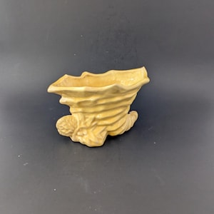 Pode incluir: Um vaso de cerâmica amarelo claro em forma de cornucópia. O vaso tem uma superfície texturizada com um design em espiral e uma borda alargada. O vaso está sobre uma superfície preta.