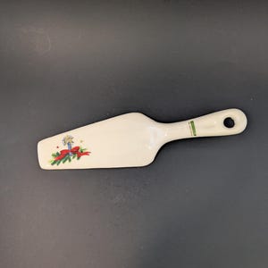 May include: A white ceramic pie server with a handle and a decorative Christmas design. The design includes a candle, red ribbon, and green pine needles. The handle has a green stripe and a hole for hanging.