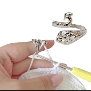 May include: A silver crochet ring with a decorative design. The ring is worn on a finger and holds yarn in place while crocheting.
