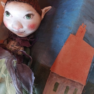 Op de afbeelding: Een fantasievolle handgemaakte pop met bruin haar, groene ogen en roze blos. Ze draagt een groene en paarse jurk met een tule rok en staat voor een geschilderde achtergrond van een huis met een rood dak.