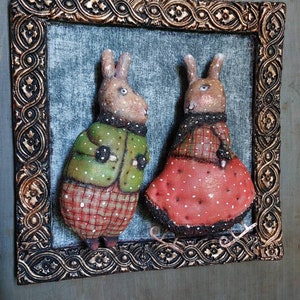 May include: Two handmade fabric rabbits in a gold frame. The rabbit on the left is wearing a green jacket with white polka dots and red and white checked trousers. The rabbit on the right is wearing a red dress with white polka dots and a black and red checked scarf.