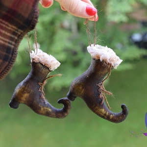 May include: Two brown felt witch shoes with pointed toes and white lace trim. The shoes are hanging from a string.