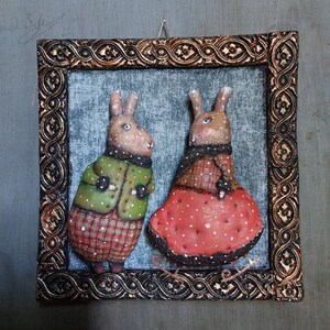 May include: A framed artwork featuring two brown rabbits wearing winter clothing. The rabbit on the left is wearing a green jacket with red and white checked accents. The rabbit on the right is wearing a red dress with white polka dots and a white trim. Both rabbits are standing on a white surface.