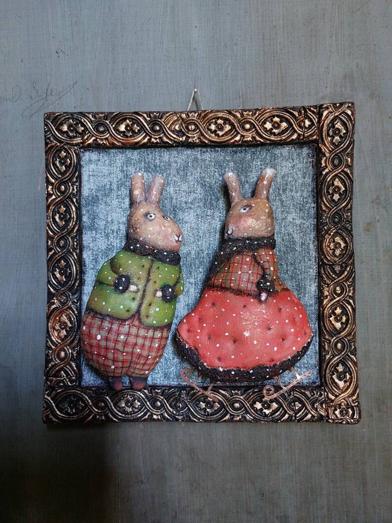 May include: A framed artwork featuring two brown rabbits wearing winter clothing. The rabbit on the left is wearing a green jacket with red and white checked accents. The rabbit on the right is wearing a red dress with white polka dots and a white trim. Both rabbits are standing on a white surface.