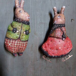 May include: Two handmade felt bunny ornaments, one wearing a green jacket and red plaid trousers, the other wearing a red dress with white polka dots and a black scarf. Both bunnies have brown fur and white eyes. The bunny in the red dress is on ice skates.