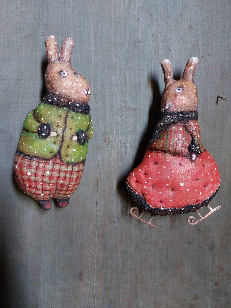 May include: Two handmade felt bunny ornaments, one wearing a green jacket and red plaid trousers, the other wearing a red dress with white polka dots and a black scarf. Both bunnies have brown fur and white eyes. The bunny in the red dress is on ice skates.