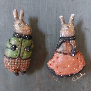 May include: Two handmade felt rabbit ornaments with painted details. One rabbit is wearing a green jacket and plaid trousers, the other is wearing a red dress with white polka dots and a plaid scarf. Both rabbits have brown fur and white accents.