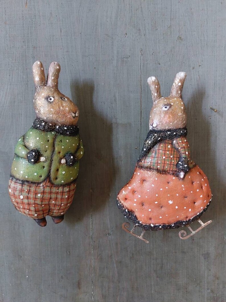 May include: Two handmade felt rabbit ornaments with painted details. One rabbit is wearing a green jacket and plaid trousers, the other is wearing a red dress with white polka dots and a plaid scarf. Both rabbits have brown fur and white accents.