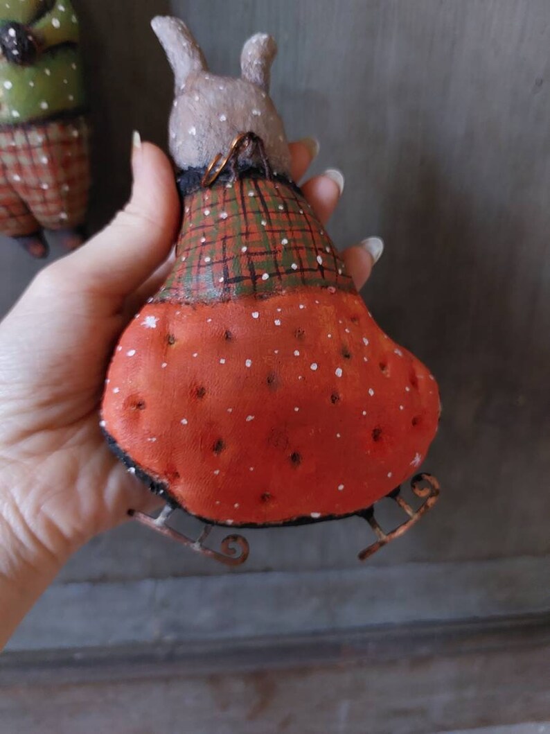 May include: A handmade felt bunny wearing a plaid and red dress with white polka dots. The bunny is on a pair of metal skis with a wire loop for hanging.