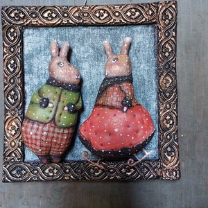 May include: Two handmade felt bunny figurines wearing winter clothes, standing on a wooden background. The bunny on the left is wearing a green jacket and red plaid trousers. The bunny on the right is wearing a red dress with white polka dots. Both bunnies are wearing black mittens.