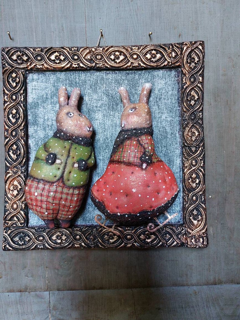 May include: Two handmade felt bunny figurines wearing winter clothes, standing on a wooden background. The bunny on the left is wearing a green jacket and red plaid trousers. The bunny on the right is wearing a red dress with white polka dots. Both bunnies are wearing black mittens.