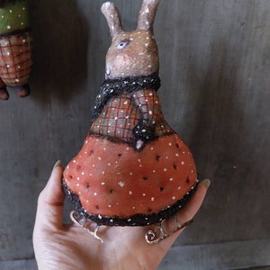 May include: A handmade brown rabbit doll wearing a red and black checked dress and a black scarf. The doll is wearing silver ice skates and is standing on a hand.