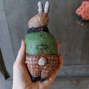 May include: A handmade plush bunny toy wearing a green and white polka dot shirt and red and white checked trousers. The bunny has a white fluffy tail and brown painted eyes.
