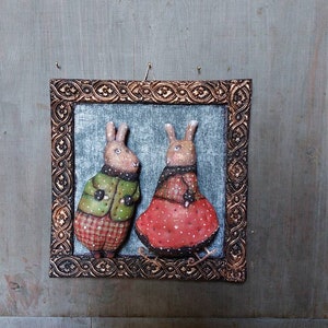 May include: A framed artwork featuring two brown rabbits wearing winter clothing. The rabbit on the left is wearing a green jacket and red plaid trousers. The rabbit on the right is wearing a red dress with white polka dots. The artwork is set against a blue background.
