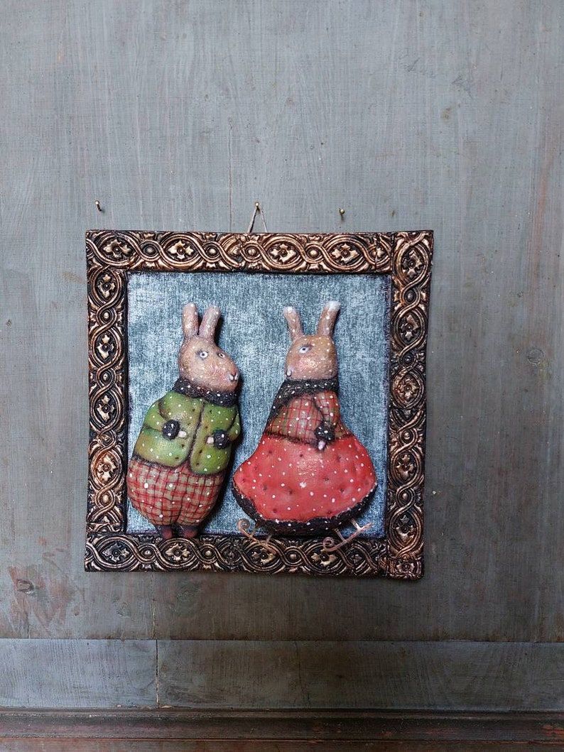 May include: A framed artwork featuring two brown rabbits wearing winter clothing. The rabbit on the left is wearing a green jacket and red plaid trousers. The rabbit on the right is wearing a red dress with white polka dots. The artwork is set against a blue background.