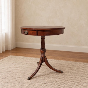May include: A round, dark brown wooden side table featuring a single drawer with a brass-toned metal pull handle. The table is supported by a central pedestal column that branches into three curved legs, each ending in a small, rounded foot.