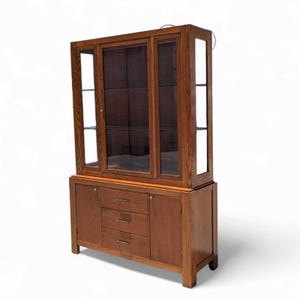 May include: A two-piece wooden display cabinet with glass doors and shelves in the upper section. The lower section has two doors and three drawers. The cabinet is a warm brown color and has a traditional design.