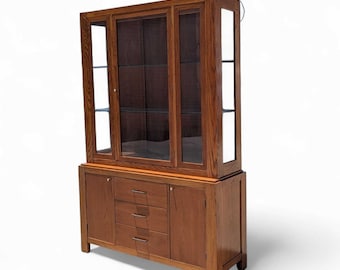 May include: A two-piece wooden display cabinet with glass doors and shelves in the upper section. The lower section has two doors and three drawers. The cabinet is a warm brown color and has a traditional design.