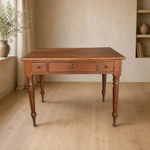 May include: A rectangular wooden table with a warm brown finish, featuring four turned legs and three front-facing drawers. The central drawer is wider than the two flanking drawers, each equipped with a small, round, dark-colored knob.