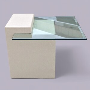 May include: A side table featuring a solid, light beige, rectangular stone base with a stepped top design. A clear, square glass tabletop is securely mounted to the upper portion of the stone base, extending horizontally to create a flat surface.