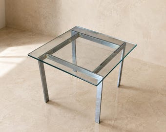May include: A square side table featuring a clear glass top supported by a metallic silver frame. The structure consists of four straight legs connected by a square metal support beam directly beneath the glass surface.