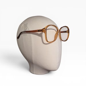 May include: Vintage oversized eyeglasses with a light brown frame and clear lenses. The glasses have a square shape and are displayed on a mannequin head. The temples are a darker brown color.