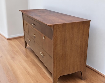 May include: A wooden dresser with a medium brown finish, featuring six drawers arranged in two columns. Each drawer is equipped with a thin, horizontal gold-toned metal handle. The piece stands on four tapered legs and has a flat rectangular top.
