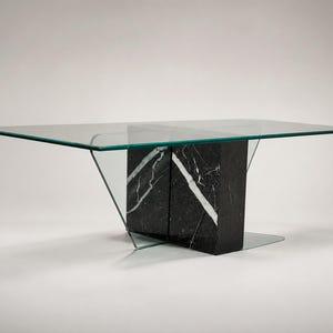 May include: A rectangular dining table featuring a clear glass top supported by a central base composed of two thick, black marble blocks with prominent white veining. The glass top rests on angled glass supports that connect to the marble base.