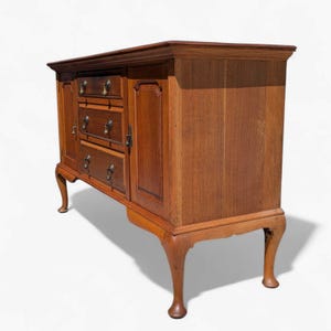 May include: A wooden cabinet with a warm brown finish. It features three drawers, two doors, and brass hardware. The cabinet has curved legs and a slightly overhanging top, showcasing a classic design.