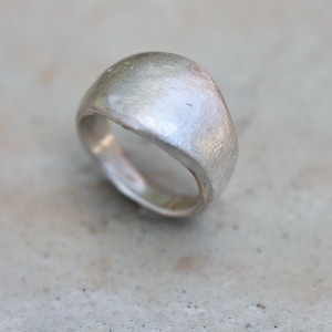May include: A silver ring with a rounded, domed top. The ring has a textured, matte finish, and the band is smooth. The ring is set against a neutral, light-colored background. The ring appears to be made of metal.