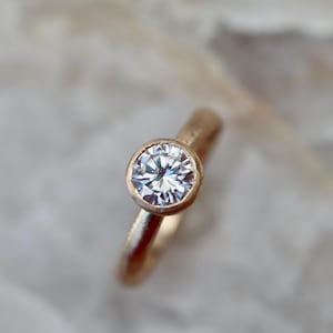 May include: A gold ring featuring a single round, clear, faceted gemstone set in a bezel setting. The band of the ring has a textured, matte finish, contrasting with the reflective, brilliant cut of the central stone.