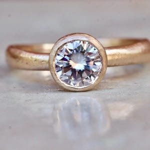 May include: A gold ring featuring a single round, clear, faceted gemstone set in a smooth bezel setting. The band has a textured, matte finish, and the center stone reflects light with multiple facets visible on its surface.
