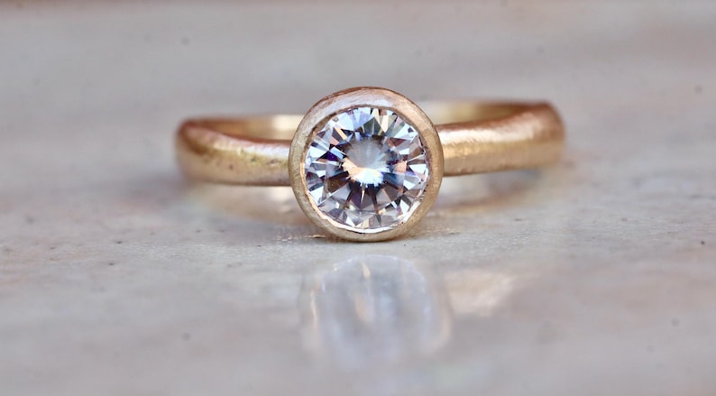May include: A gold ring featuring a single round, clear, faceted gemstone set in a smooth bezel setting. The band has a textured, matte finish, and the center stone reflects light with multiple facets visible on its surface.