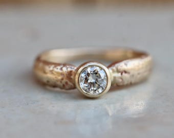 May include: A gold ring with a textured band and a round, clear diamond set in a bezel. The ring is set against a neutral background, highlighting the sparkle of the diamond.
