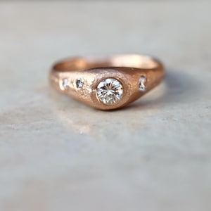 May include: A rose gold ring with a central, round, clear gemstone and smaller accent stones. The ring has a textured surface and a slightly irregular shape, giving it a unique, handcrafted appearance. This ring is a piece of jewelry.