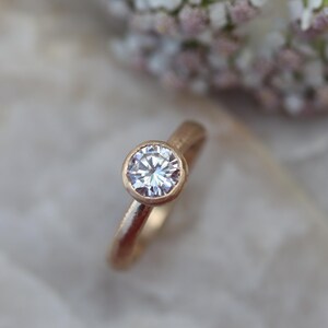 May include: A gold ring featuring a single round, clear, faceted gemstone set in a bezel setting. The band of the ring has a textured, matte finish, contrasting with the reflective, brilliant cut of the central stone.