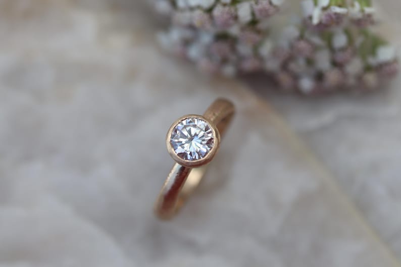May include: A gold ring featuring a single round, clear, faceted gemstone set in a bezel setting. The band of the ring has a textured, matte finish, contrasting with the reflective, brilliant cut of the central stone.