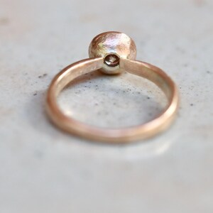 May include: A rose gold ring featuring a smooth, rounded band and a single, small, clear, round-cut gemstone set in a bezel setting. The metal surface has a soft, warm, metallic luster, and the ring is positioned to show the side profile.